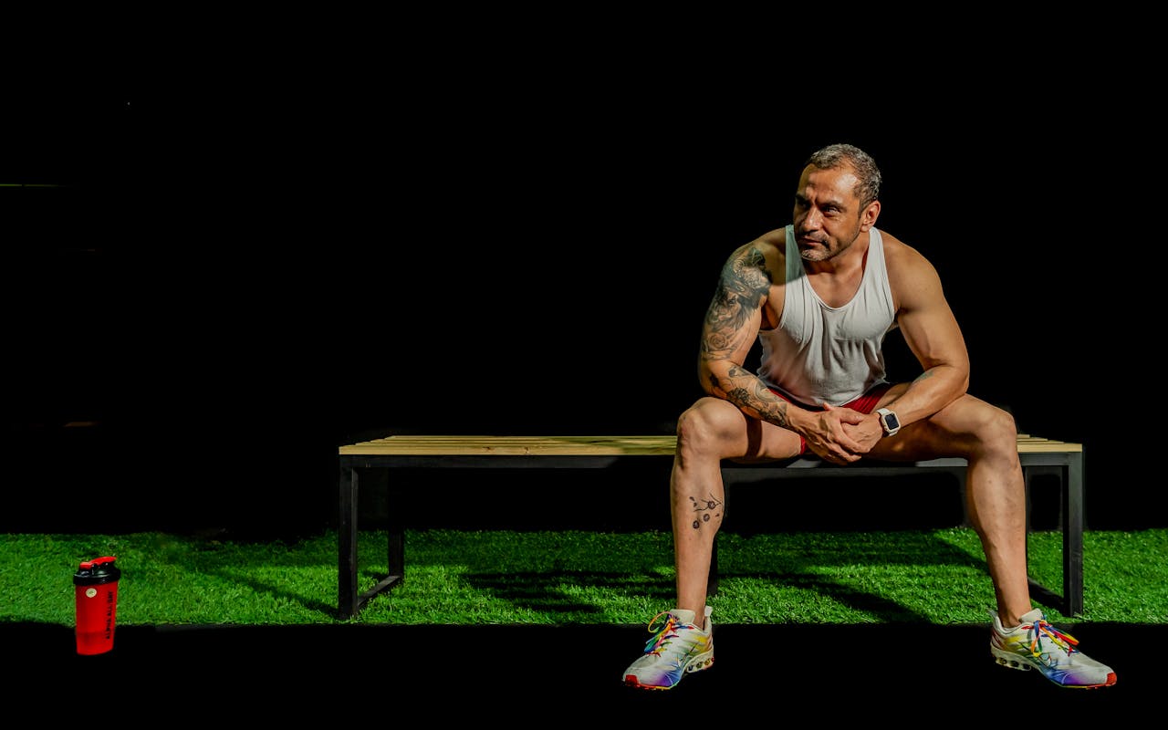 A muscular, tattooed man seated on a bench in a gym, depicting modern fitness lifestyle.